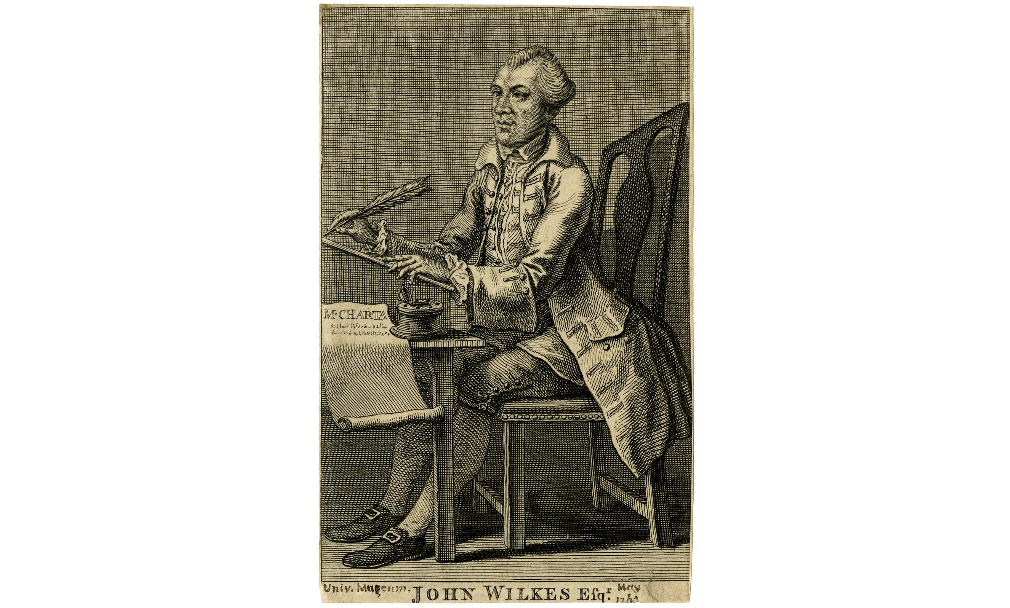 John Wilkes is sat on a chair at a desk that has a writing slope on top. He is holding a feather quill in his right hand. On the desk is an unrolled piece of paper with writing on it and an ink pot. Wilkes is wearing a long coat, breeches, and shoes with a buckle.