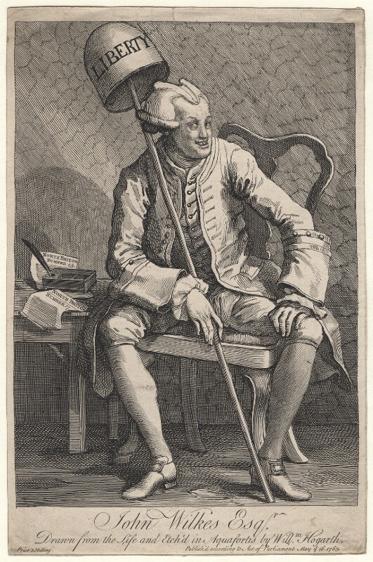 A caricature of a white man wearing a light-coloured wig. He is sat on a chair next to a table. The table has two pieces of paper, an inkwell and a quill. The man is holding a pole with a bell on top of it with the writing 'LIBERTY' in his right hand. His left hand is resting on his left knee. He is wearing a jacket, breeches, and shoes.