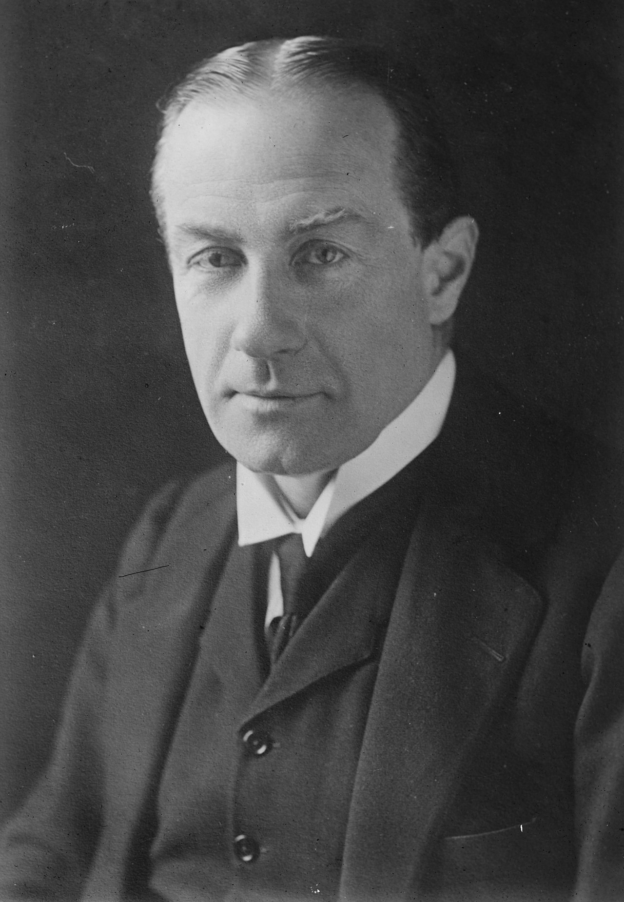 A black and white photograph of a white man. His torso and head are in frame. He has thinning dark hair. He is clean shaven. He is wearing a dark three piece suit with white shirt and a dark tie.