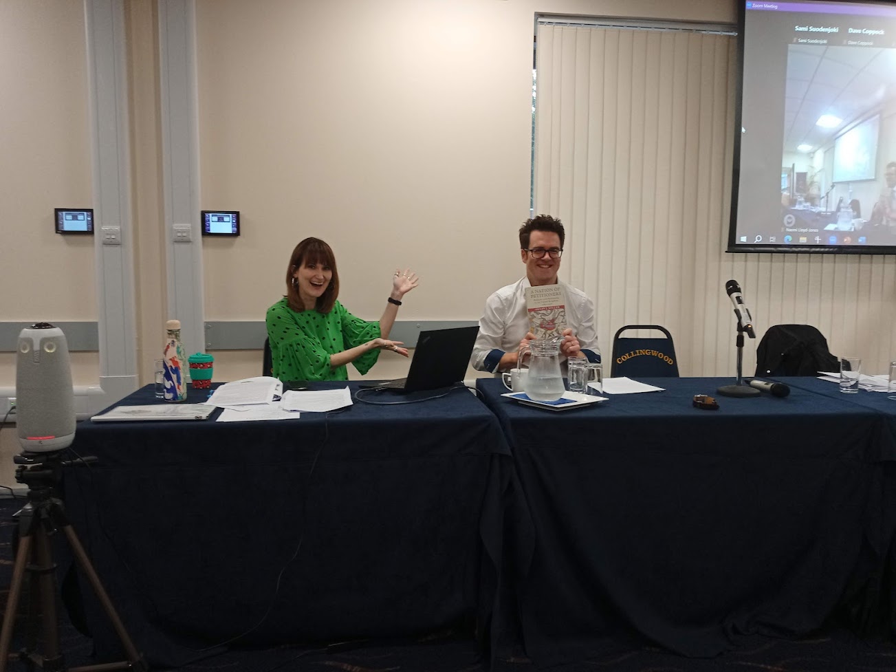 A white woman and a white man sat next to each other. The woman is wearing a green dress. She has a fringe. She is pointing towards the man. The man has short dark hair, glasses and is wearing a white shirt. He is smiling and holding a book.