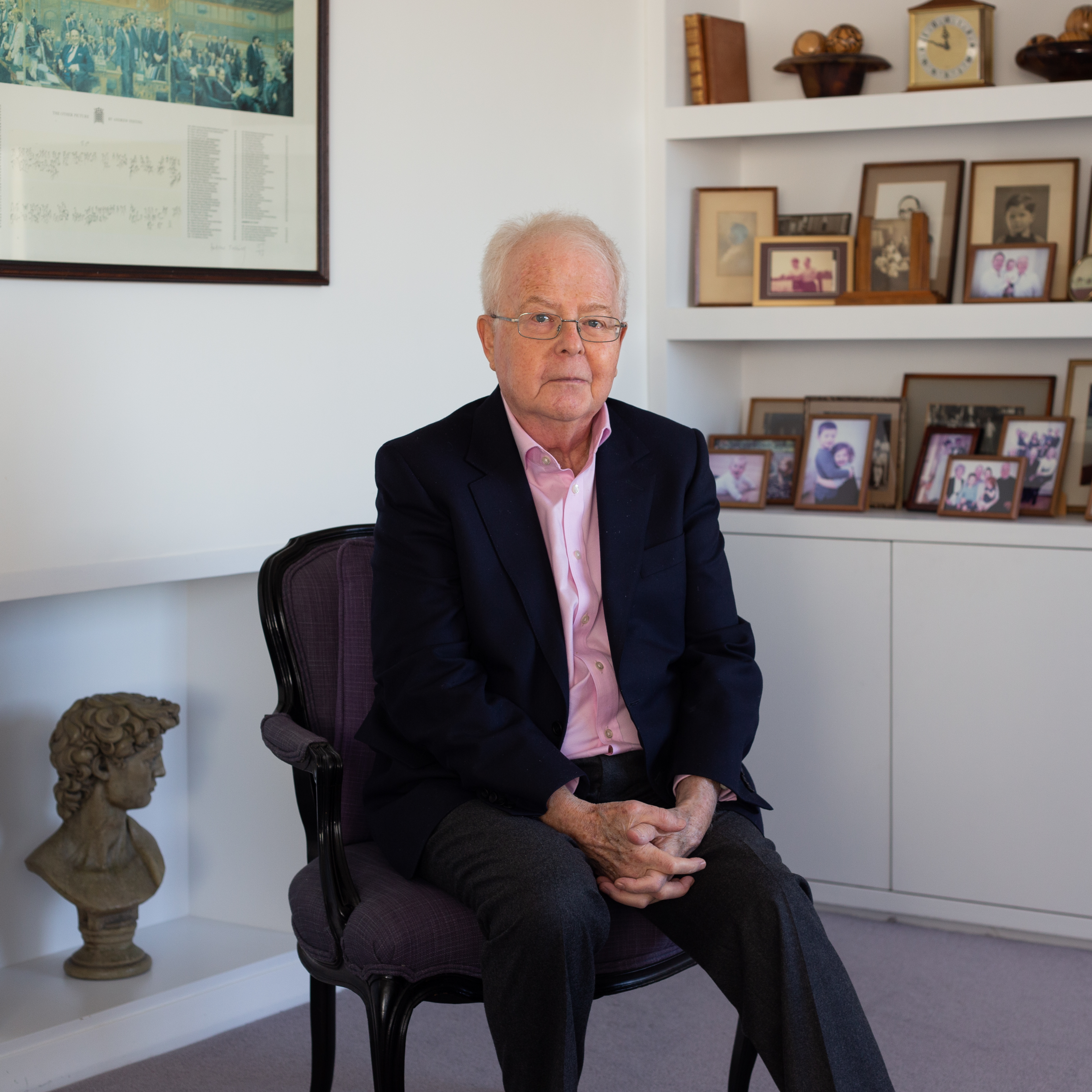 Portrait of David Sumberg. He is say on a purple upholstered chair with his hands clasped on in lap. He wears a light pink shirt, grey trousers and navy suit jacket. Behind David is a bookshelf covered with many family photographs. 
