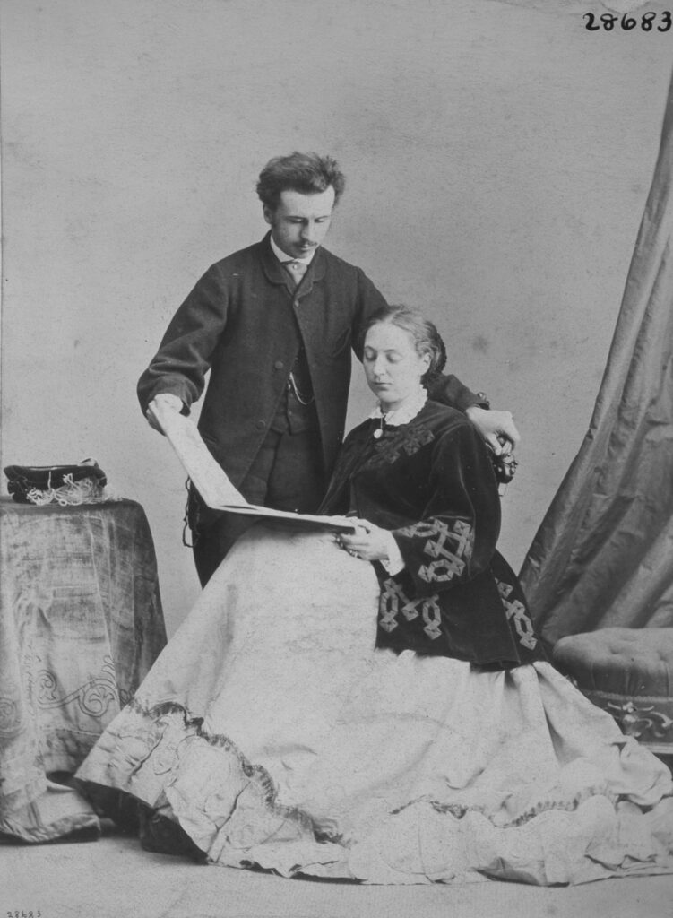 Albumen print, a black and white full-length photographic portrait of John Russell and Katharine Louisa Russell, Viscount and Viscountess Amberley. Sitting down to the right centre is the Viscountess. She is wearing a light wide dress with a black coat with an angular knotted design across the coat, her hair is swept back into a bun. She is holding a wide book open reading, whilst the Viscount is holding the right page with his hand, standing up to her right. He is wearing a three piece black wool suit, with a white shirt and no tie. He has a neat moustache and full black hair. His left hand is holding the back of the chair the Viscountess is sitting on. There is a table with a tablecloth to the left and a curtain and stool to the right of the picture. 