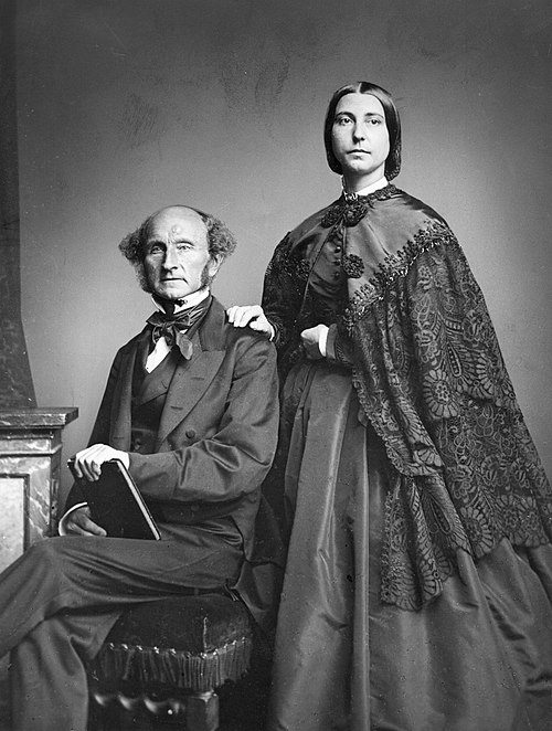 A black and white photograph portrait of John Stuart Mill and Helen Taylor. John, sitting down on a chair, is wearing a black three-piece suit with a white high collar shirt and black necktie. He has frizzy curly hair on the sides of his head and bald on top, and has long sideburns. He is holding a book in his hands and lap. To the right, Helen is standing, wearing a black wide dress and a lace detailed black cape. Her hair is middle parted black hair which is done up shortly behind her head.
