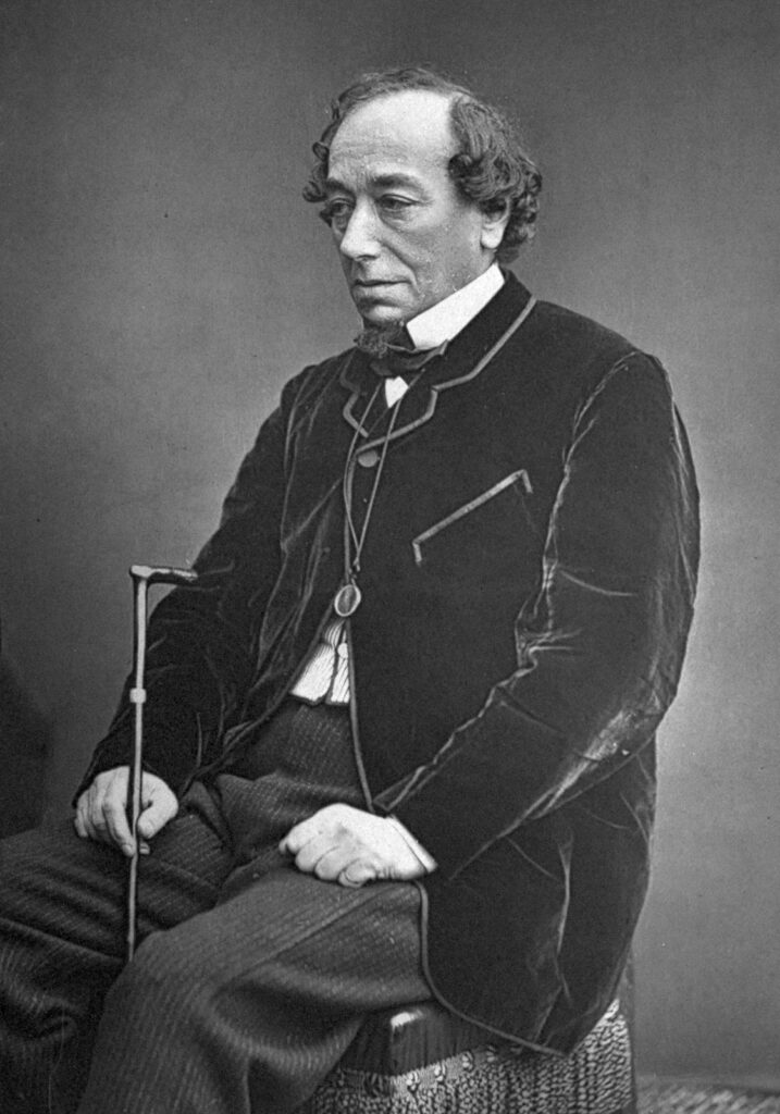 A black and white photograph portrait of Benjamin Disraeli. Sitting down on a stool, he is wearing grey pinstriped trousers, a black suit jacket with pronounced bordering, a white shirt and black necktie. In his right hand and between his legs he holds a walking stick. He is clean shaven with combed receding wavy hair.