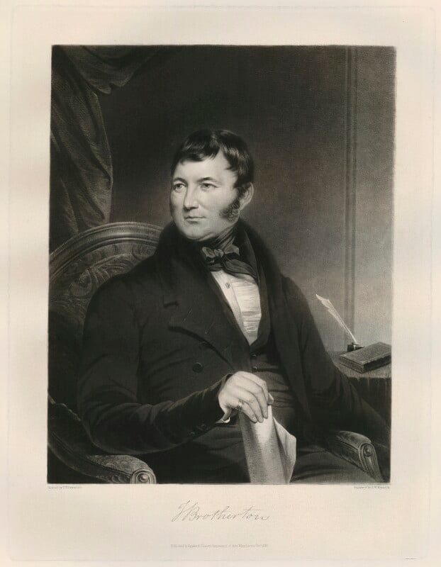 A half length black and white portrait of Joseph Brotherton. Sitting in a high backed chair, he is wearing a dark suit jacket, a dark waistcoat, white shirt and high black necktie. He is holding rolled parchment from the end in his right hand. Looking slightly to the left of the image he has short black hair with long sideburns. To his right behind him on a table is a closed book with a quill in an inkwell.