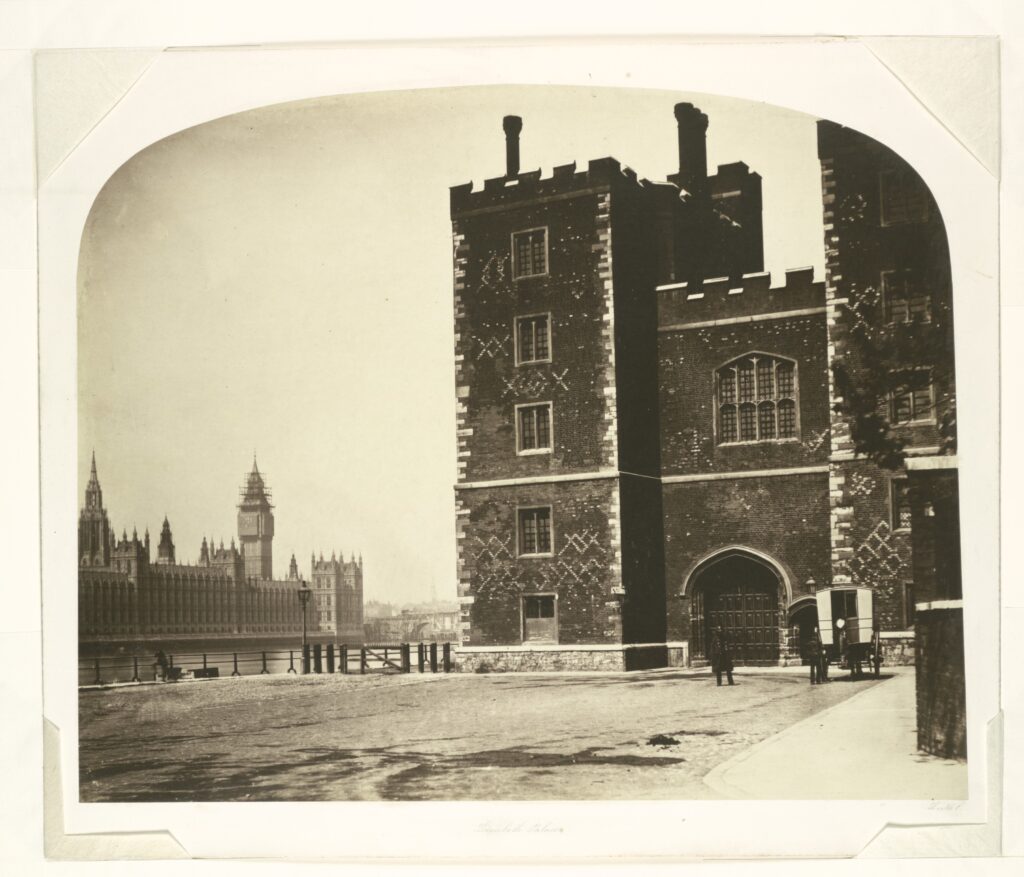 A black and white photograph of Lambeth Palace on yellowed paper. From the centre to the right, with a large drive in front of it stands Lambeth palace, with a dark painted exterior walls, with two low square towers with crenelated tops. To the left and in the background on the other side of the river, stands the palace of Westminster, with the top of big ben under construction.