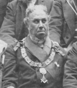 A cropped black and white photograph of Peter McLagan in full masonic attire. He has a dark complexion, a greying beard under his neck but a clean shaven face, and short greying hair. 