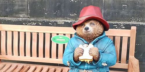 A photograph of a statue of paddington bear sitting on a wooden bench with a green plaque on it. He is a small brown bear with a red hat and blue duffel coat. He is holding a marmalade sandwich with a bite taken out of it. 
