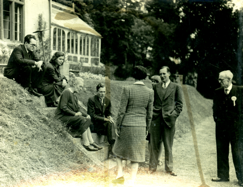 A black and white faded picture of the Foot family outside. To the left four of them are sitting on steps on a grassy bank. Four to the right are standing on a path. The man to the furthest right is wearing a dark suit with a light rosette pinned to his lapel.