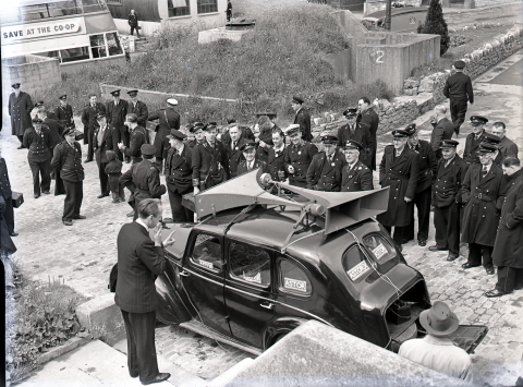 A black and white photograph of a a man speaking into a handheld microphone for a loudspeaker attached to the top of a black car which has 'Astor' posters in the windows. Surrounding the car is a group of uniformed men listening to the speaker.