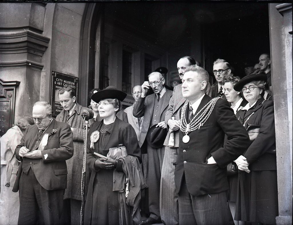 A black and white photograph on the steps of the city museum and art galleries, Plymouth. A collection of men and women are standing on the steps looking out. A women in dark dress, leather gloves, dark bonnet and a rosette pinned to her lapel, is speaking into a microphone in a stand. 